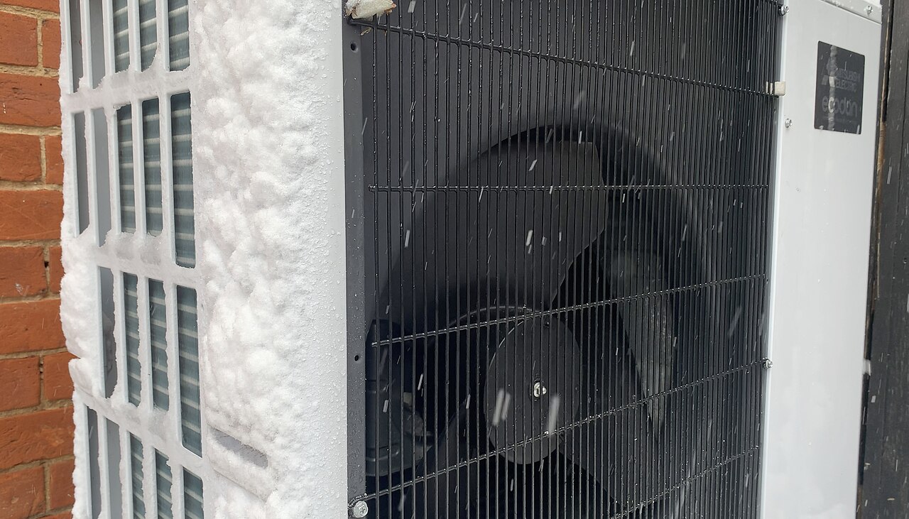 A Mitsubishi heat pump outdoor unit with snow packed on the coil beside a brick wall